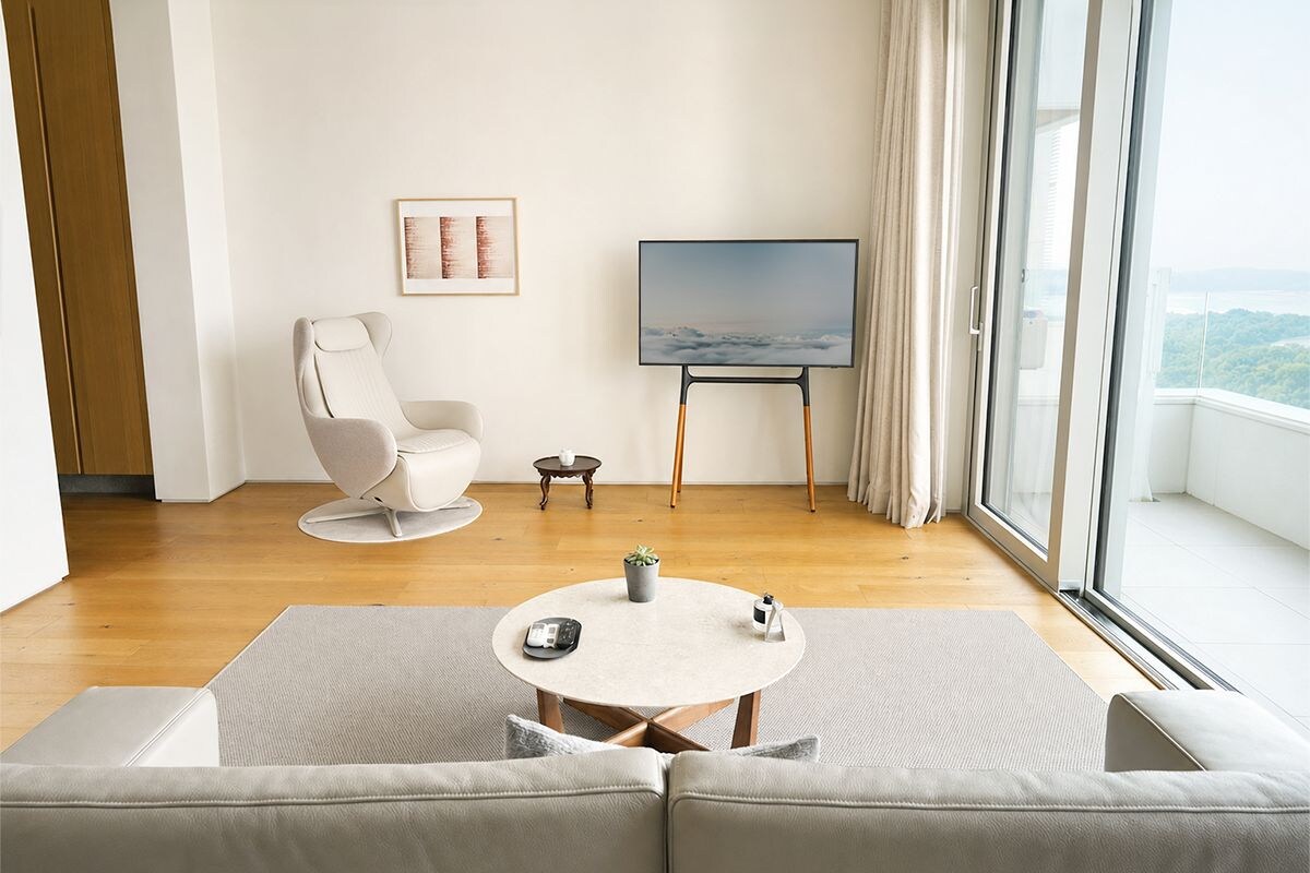 Ein modernes Wohnzimmer mit einer Couch, einem kleinen Tisch, einem Sessel, einem Fernseher auf einem Schrank und Blick durch große Fenster.