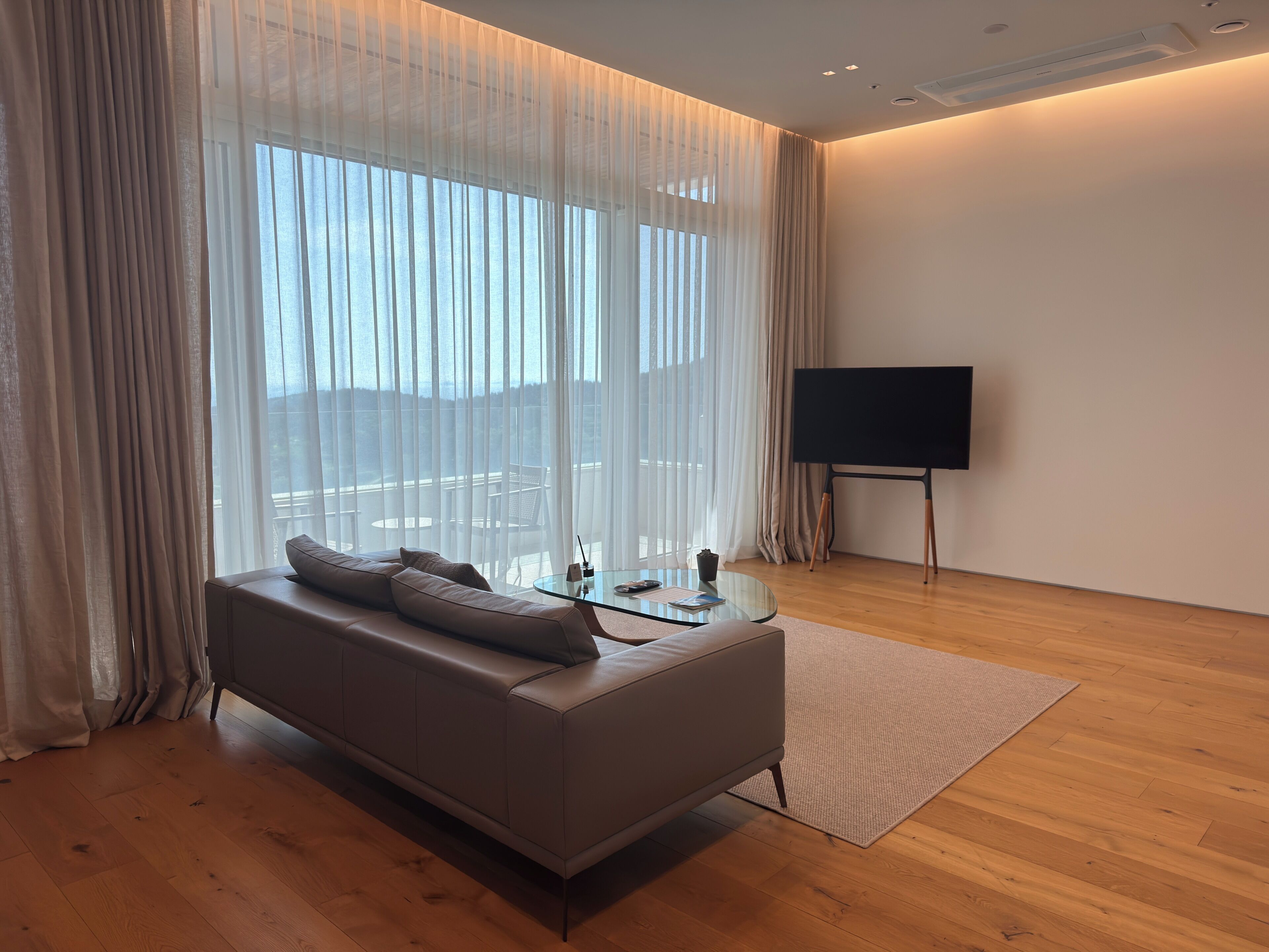 Een moderne woonkamer met een bank, een glazen salontafel, een flatscreen televisie en dunne gordijnen.