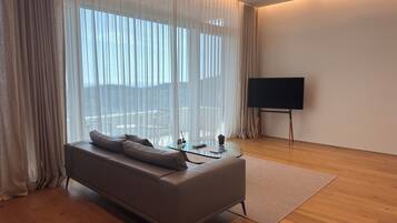 Een moderne woonkamer met een bank, een glazen salontafel, een flatscreen televisie en dunne gordijnen.