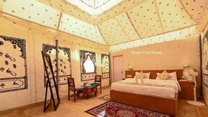 A bedroom with a bed, a desk, and chairs, decorated with patterned wallpaper and a large rug.