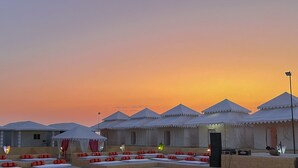 A row of white tents with a flat roof, arranged in a row. Each tent has a small porch area with a bench and a table.