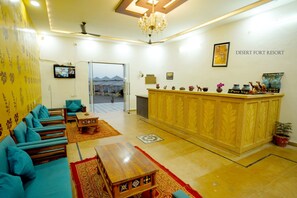 A reception area with a reception desk, seating area with sofas, a chandelier, and a view of the desert through the glass door. - Desert Fort Resort (Jaisalmer)