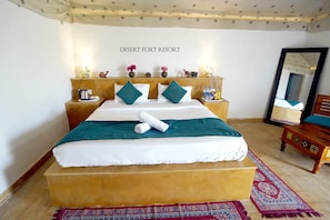A hotel room with a large bed, a headboard with decorative items, a nightstand with a lamp, and a mirror on the wall. - Desert Fort Resort (Jaisalmer)