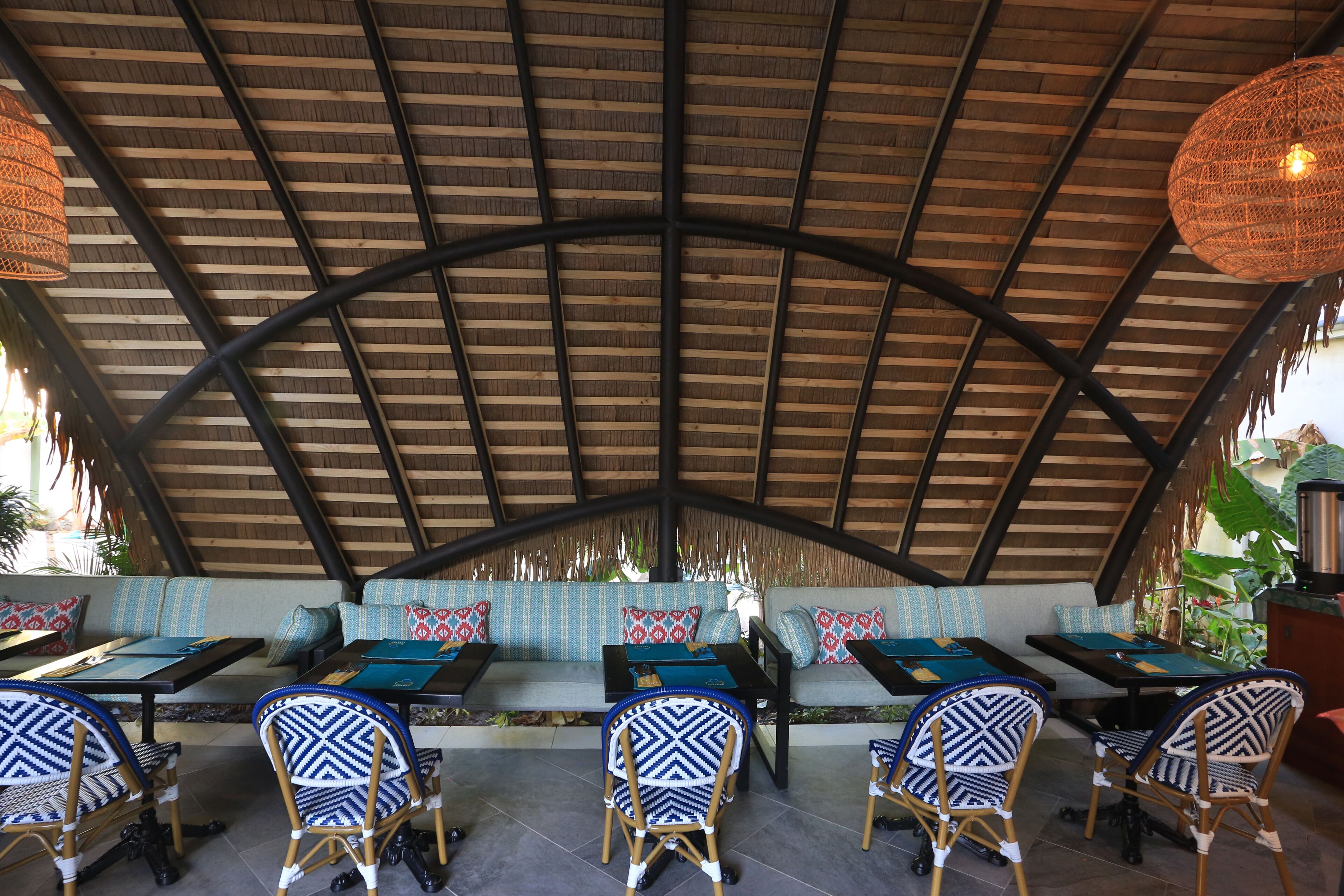 A covered outdoor seating area with tables and chairs, a thatched roof, and a sofa with patterned cushions.