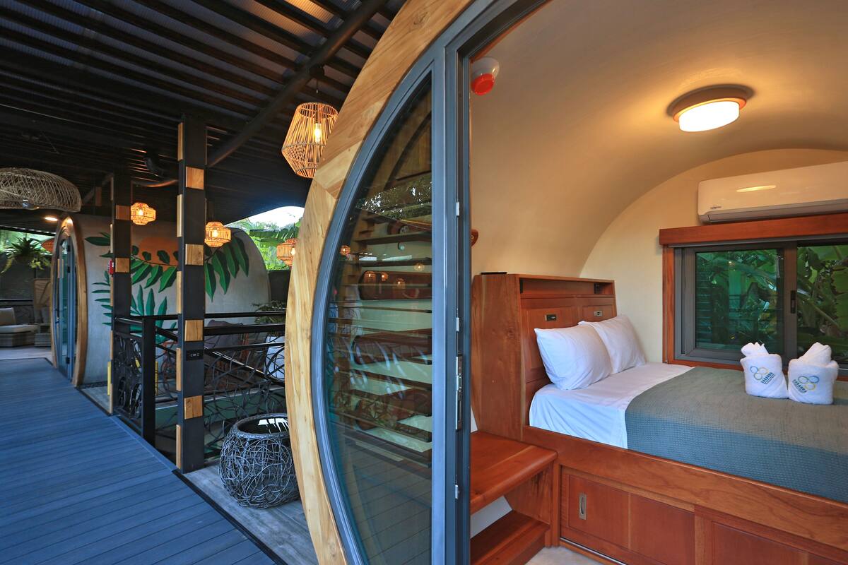 Una habitación con un muro de cristal curvo, una cama de madera y un pequeño rincón de estar.