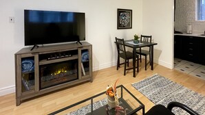 Ein Wohnzimmer mit Flachbildfernseher, Kamin, Esstisch mit Stühlen und einer durch einen offenen Durchgang sichtbaren Küche.