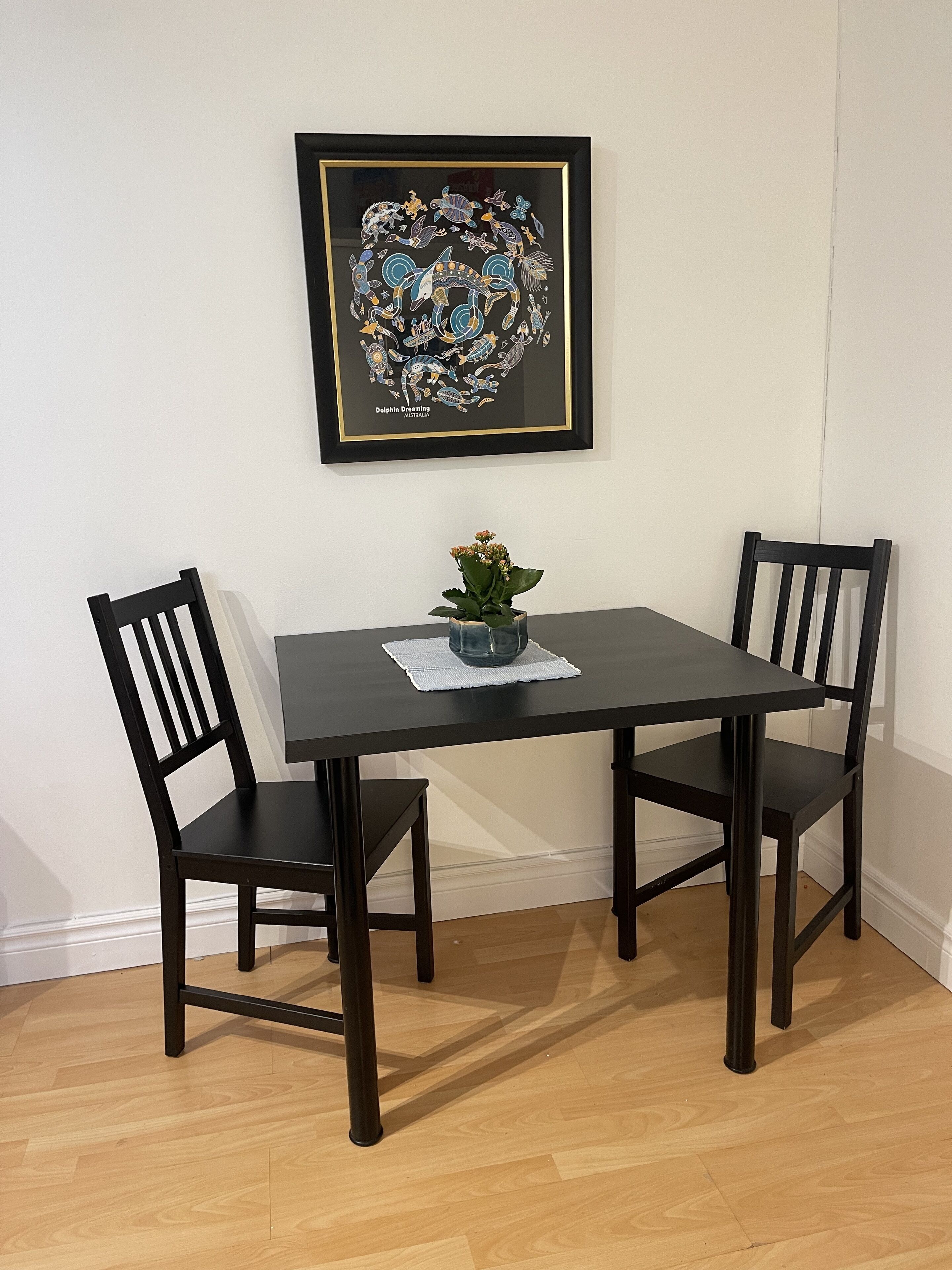 Une table à manger avec deux chaises, une œuvre d’art encadrée au-dessus, et une petite plante posée sur la table.