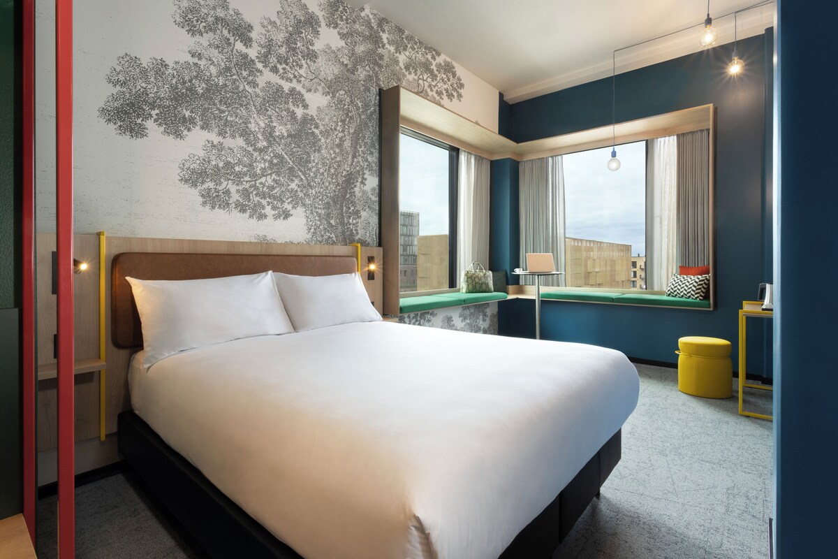Habitación de hotel con una cama grande, una ventana con vistas a edificios, una mesita con una lámpara y un taburete amarillo.