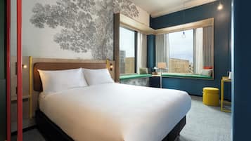 Een hotelkamer met een groot bed, een raam met uitzicht op gebouwen, een kleine tafel met een lamp en een gele stoel.