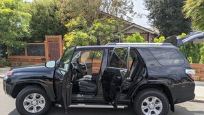 Ein schwarzer SUV steht auf einer Auffahrt mit geöffneten Türen, umgeben von Grünpflanzen und einem Holzzaun.