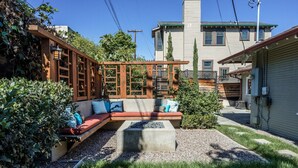 Ein Garten mit einer Holzterrasse, einer Beton-Feuerstelle und einem roten Sofa mit Kissen.