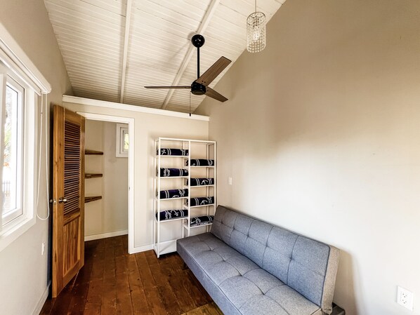 Un pasillo con una puerta de madera, un estante blanco con cojines estampados, un banco tapizado gris y un ventilador de techo.