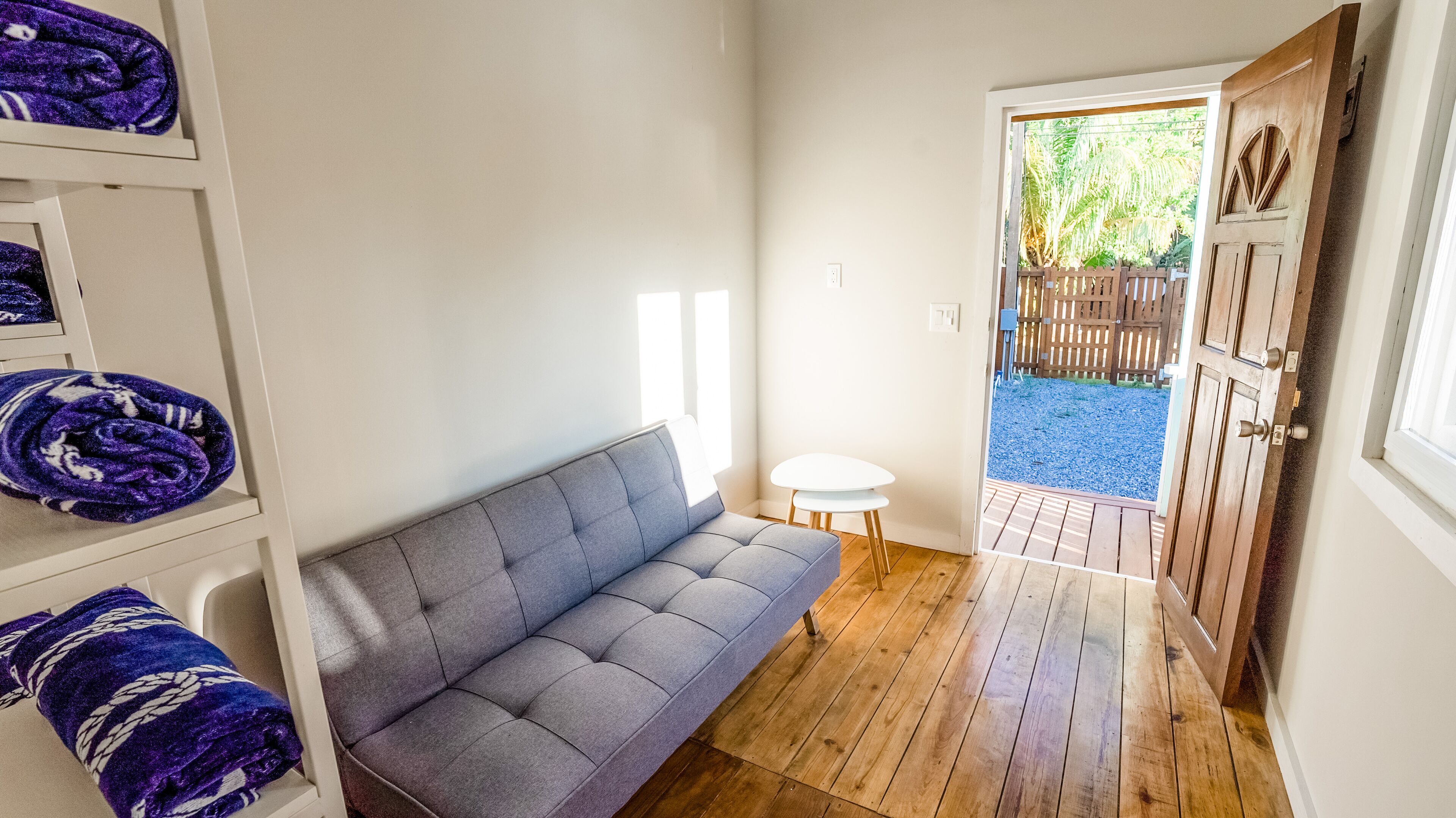 A room with a grey sofa, a small white table, and a wooden door leading to an outdoor area with a gravel path and wooden fence.