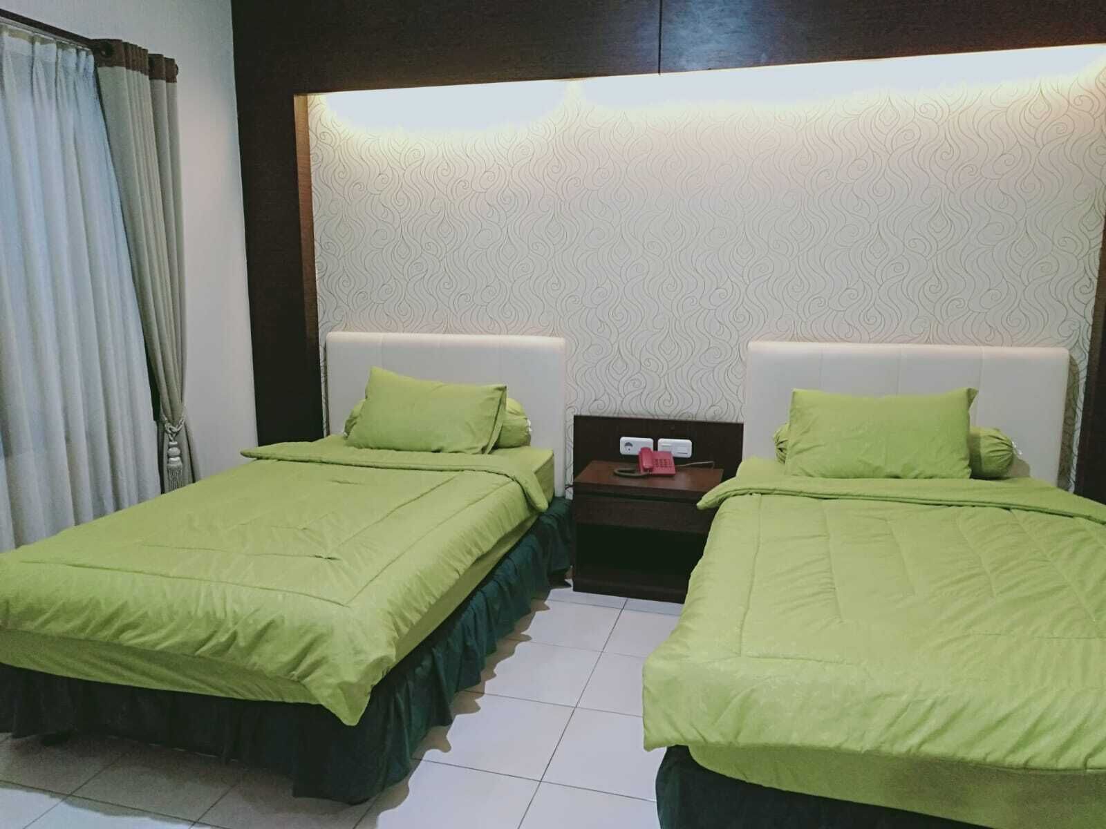 A hotel room with two beds, each with green bedding, a nightstand with a phone, and a patterned wallpaper.