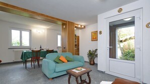 Ein Wohnzimmer mit einem petrolfarbenen Sofa, einem Holztisch und einem braunen Kissen. Es gibt einen Essbereich mit einem grünen Tisch und Stühlen. Eine Topfpflanze steht auf dem Boden, und eine Tür führt zu einem Außenbereich.