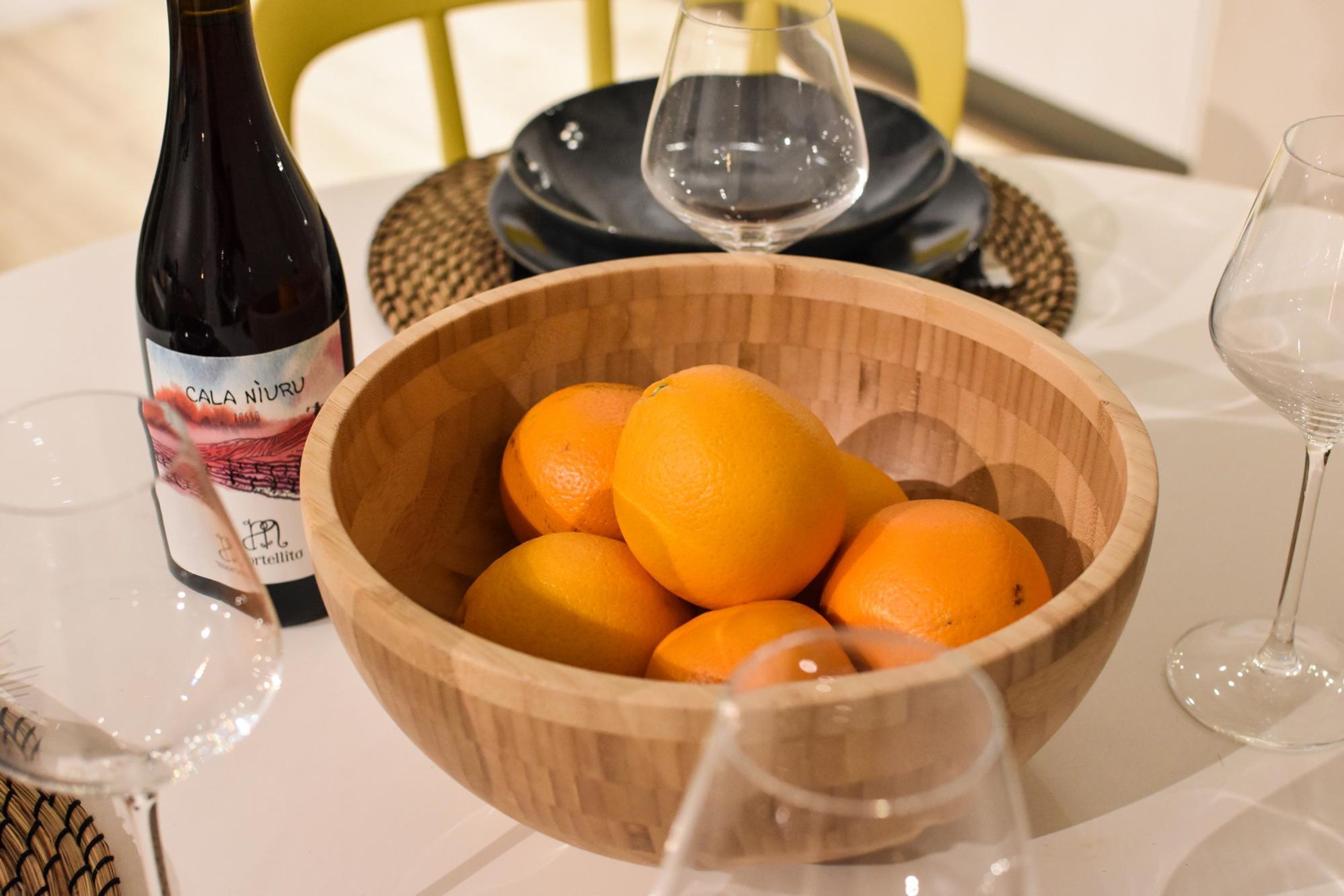 Un saladier en bois rempli d’oranges, une bouteille de vin et des verres à vin sur une table.
