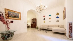 A spacious living room with a chandelier, a sofa, a dining area, and a hallway with a painting and a vase of flowers. - Palazzo Dello Spagnolo Suite (Naples)