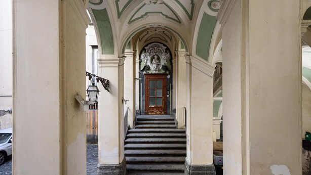 A corridor with a wooden door at the end, flanked by columns and a decorative ceiling. - Palazzo Dello Spagnolo Suite (Naples)