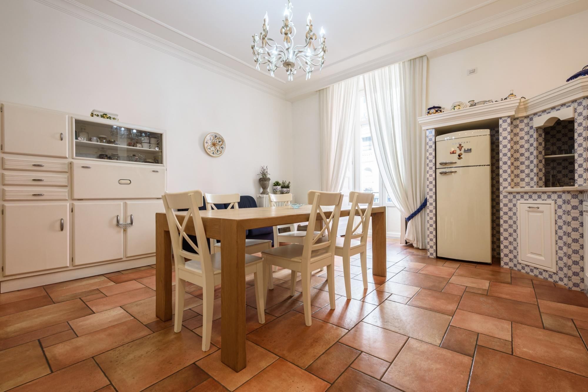 Zona pranzo con tavolo e sedie in legno, un lampadario e una cucina con carta da parati decorata e un frigorifero.
