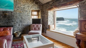Ein gemütliches Zimmer mit Steinwänden, einem großen Fenster mit Meerblick, einem komfortablen rosa Sofa und einem Glastisch.