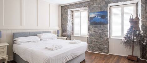 Een slaapkamer met een groot bed, witte beddengoed, een stenen muur en een raam met uitzicht op de bergen.