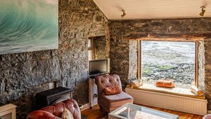 Ein gemütliches Wohnzimmer mit einer Steinwand, einem großen Gemälde mit Wellenmotiven, einer Holzbalkendecke mit Steuerrad und Blick ins Freie durch ein Fenster.