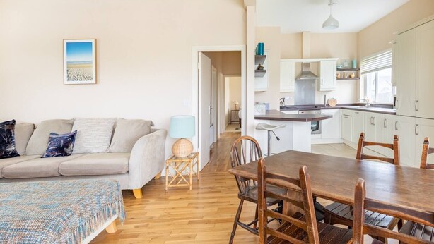 Un vaste espace de vie comprenant un canapé, une table basse et une table à manger avec des chaises. On aperçoit au fond une cuisine avec des meubles blancs et une cuisinière.