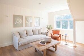 A modern living room with a beige sofa, a wooden coffee table, and a small orange armchair. There are three framed artworks on the wall and a window with a view of greenery outside. - The Links 10, Amazing Location- Food, Drinks, Surf, & Golf! - 10 (Lahinch)