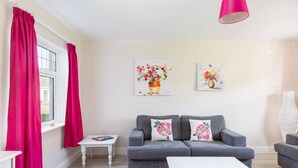 Ein Wohnzimmer mit einem grauen Sofa, einem weißen Couchtisch und einem kleinen weißen Beistelltisch. An der Wand hängen zwei Blumenbilder und eine rosa Lampe.