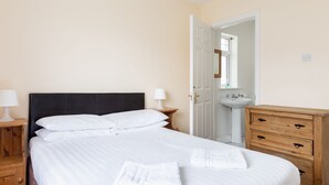 Una cama bien hecha con sábanas blancas y dos toallas, una mesita de noche con lámpara, un aparador de madera con cajones, y un baño blanco con lavamanos y espejo.