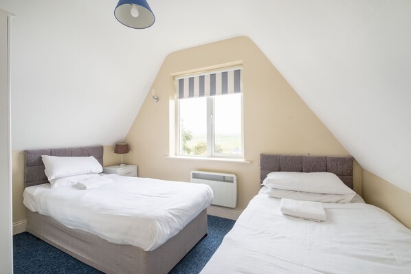 A bedroom with two beds, a window with a striped blind, a bedside table with a lamp, and a radiator. - The Links 04, Amazing Location- Food, Drinks, Surf, & Golf!-4 (Lahinch)