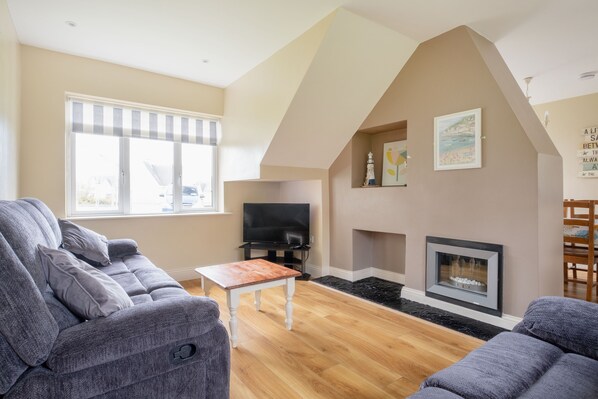 A living room with a grey sofa, a wooden coffee table, a flat-screen TV, a fireplace, and a large window with blinds. - The Links 04, Amazing Location- Food, Drinks, Surf, & Golf!-4 (Lahinch)