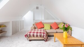 A cozy loft bedroom with a sloped ceiling, a beige sofa with colorful cushions, a plaid throw blanket, and a wooden nightstand with a vase of flowers. - The Play School, The Play School (Miltown Malbay)