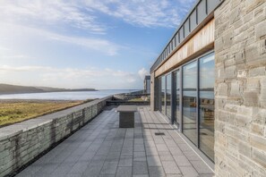 A coastal building with large glass windows, a stone wall, and a paved walkway leading to the sea. - Cregg Beach - Luxury in Lahinch! (Lahinch)