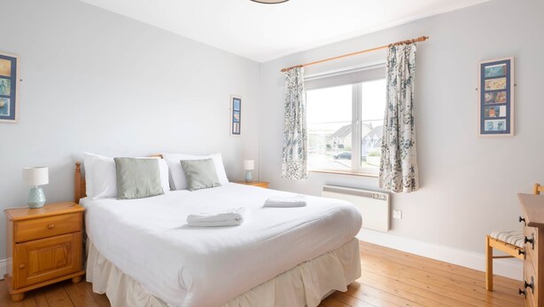 A bedroom with a large bed, wooden floor, a nightstand with a lamp, and a window with curtains. - Summer Cove 10 (900m to Lahinch Beach) (Lahinch)