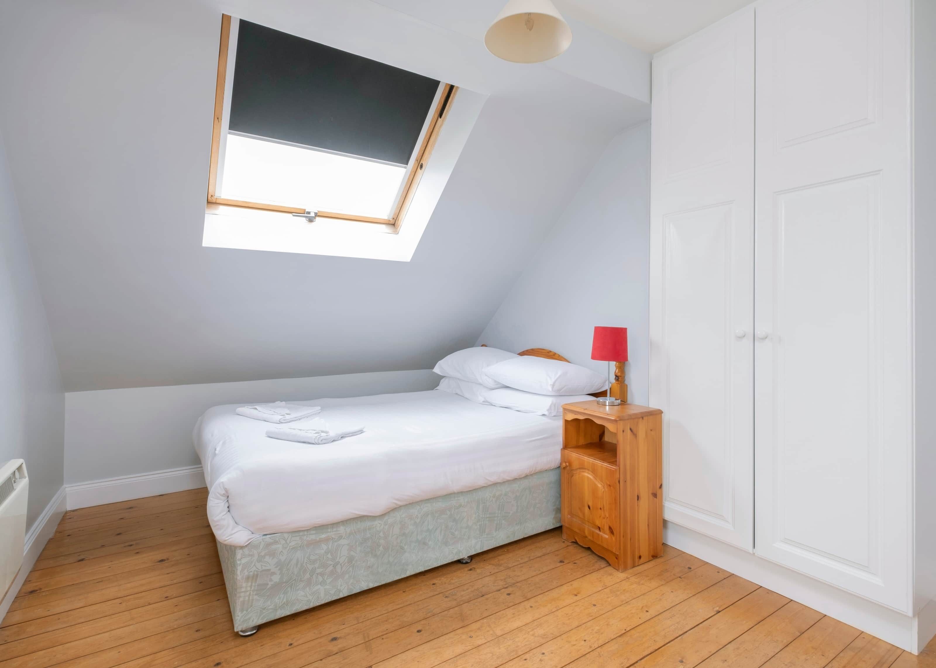 Ein Schlafzimmer mit Oberlichter, einem Bett mit weißer Bettwäsche, einem Nachttisch aus Holz mit einer roten Lampe und einem weißen Kleiderschrank.