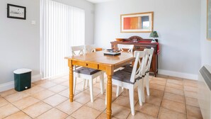A dining area with a wooden table and white chairs, a window with blinds, a framed picture on the wall, and a small trash bin. - Summer Cove 10 (900m to Lahinch Beach) (Lahinch)