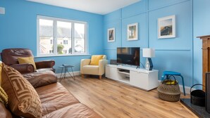 Un salon aux murs bleus, avec un canapé en cuir marron, un fauteuil blanc, un téléviseur et une petite table.