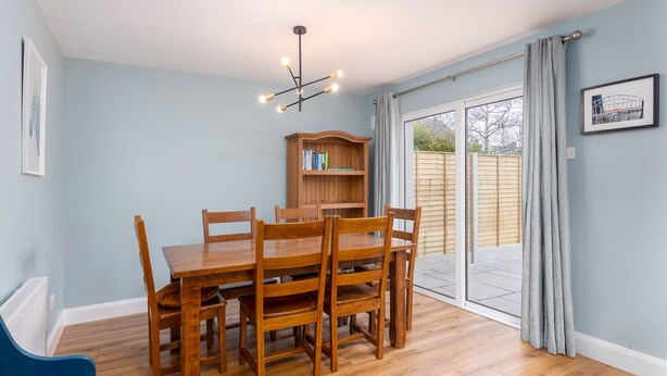 Un coin repas avec une table et des chaises en bois, un luminaire au plafond, une bibliothèque et une vue sur l’extérieur, accessible par une porte vitrée.