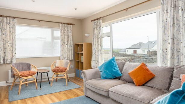 Een gezellige woonkamer met een bank, twee fauteuils, een bijzettafeltje en een boekenkast. Grote ramen met gordijnen bieden uitzicht op het omliggende landschap.