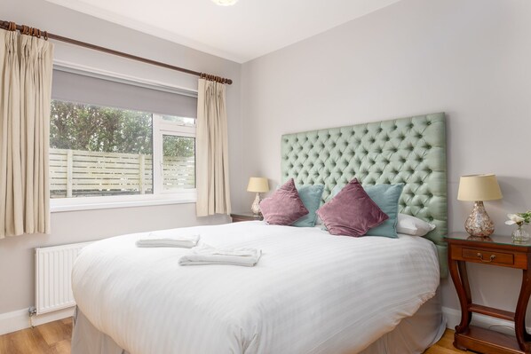 A neatly made bed with white linens and two pillows, a green tufted headboard, a bedside table with a lamp, and a window with curtains.