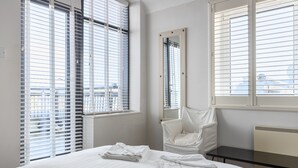 Une chambre d’hôtel avec un lit, une chaise et une table. Il y a des fenêtres avec des stores et un miroir au mur.