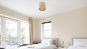 Ein Schlafzimmer mit zwei Betten, einem Korbsessel, einem Nachttisch mit Lampe und einem Fenster mit Vorhängen.