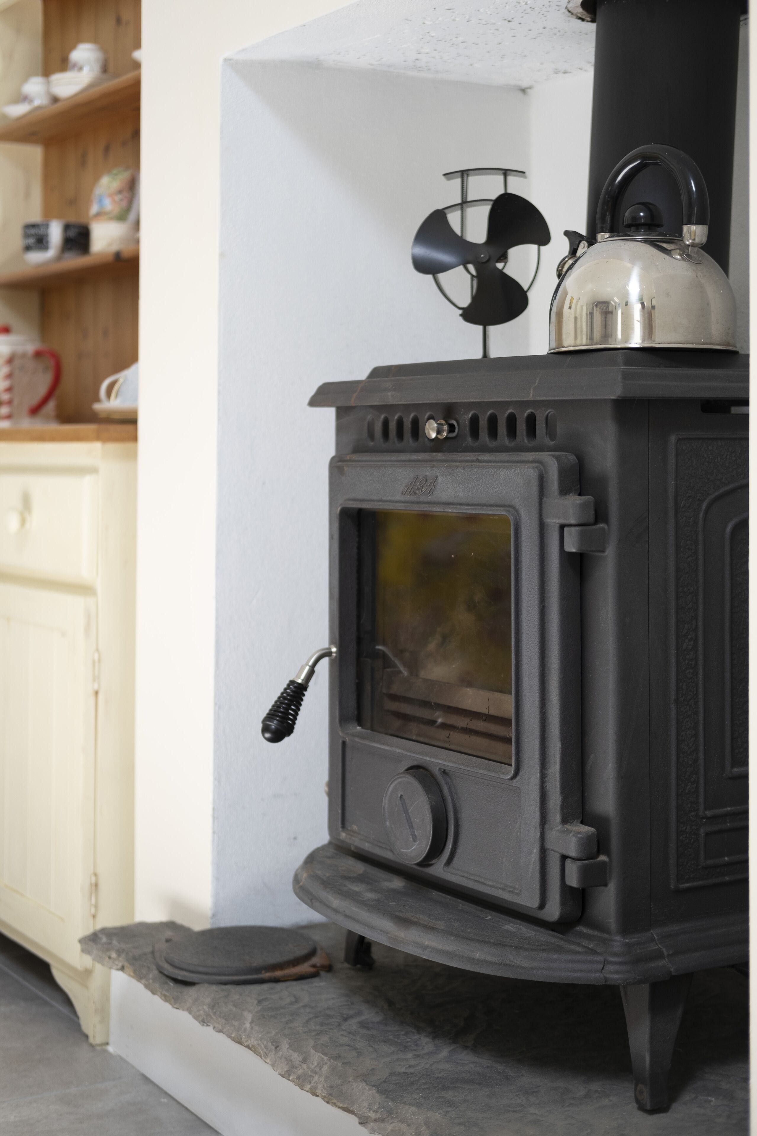 Un poêle à bois noir, avec une bouilloire posée sur le dessus, un ventilateur et une fenêtre.