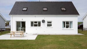 A white single-story house with a black roof, a small patio with a wooden table, and a grassy yard. - Clochan Na Mara 15 (In Liscannor Village) (County Clare)