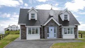 A two-story house with a stone facade, a grey roof, and a front door with a small porch. - Clochan Na Mara 15 (In Liscannor Village) (County Clare)