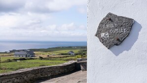 Un mur de pierre sur lequel est fixée une plaque portant l’inscription « Kneely Cottage », et une vue sur un paysage côtier avec des maisons et des champs.
