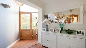 Une pièce avec une porte en bois donnant sur l’extérieur, un télescope posé sur un trépied, une commode blanche avec des tiroirs, un miroir et un vase contenant des fleurs séchées.