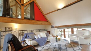 Ein gemütliches Loft-Wohnzimmer mit hoher Decke, freiliegenden Holzbalken, einem weißen Sofa, einem weißen Sessel und einem runden Couchtisch.