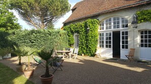 A house with a gravel driveway, a patio with a table and chairs, and a large tree in the foreground.